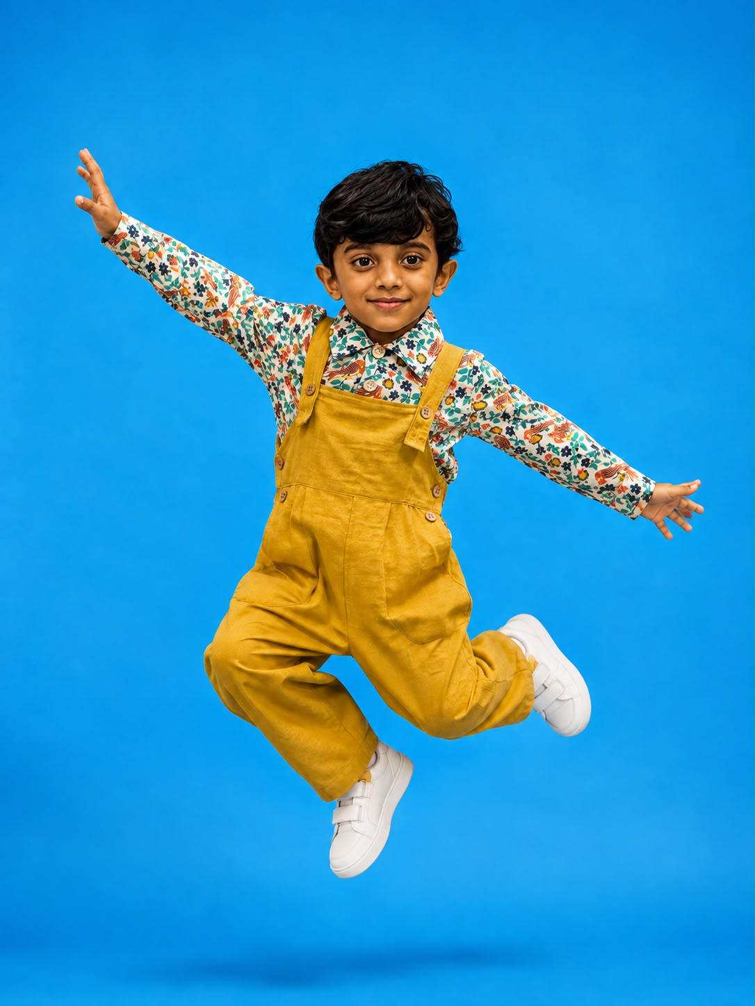 Boy wearing mustard yellow khadi dungaree with floral print shirt, jumping mid-air against a blue background.