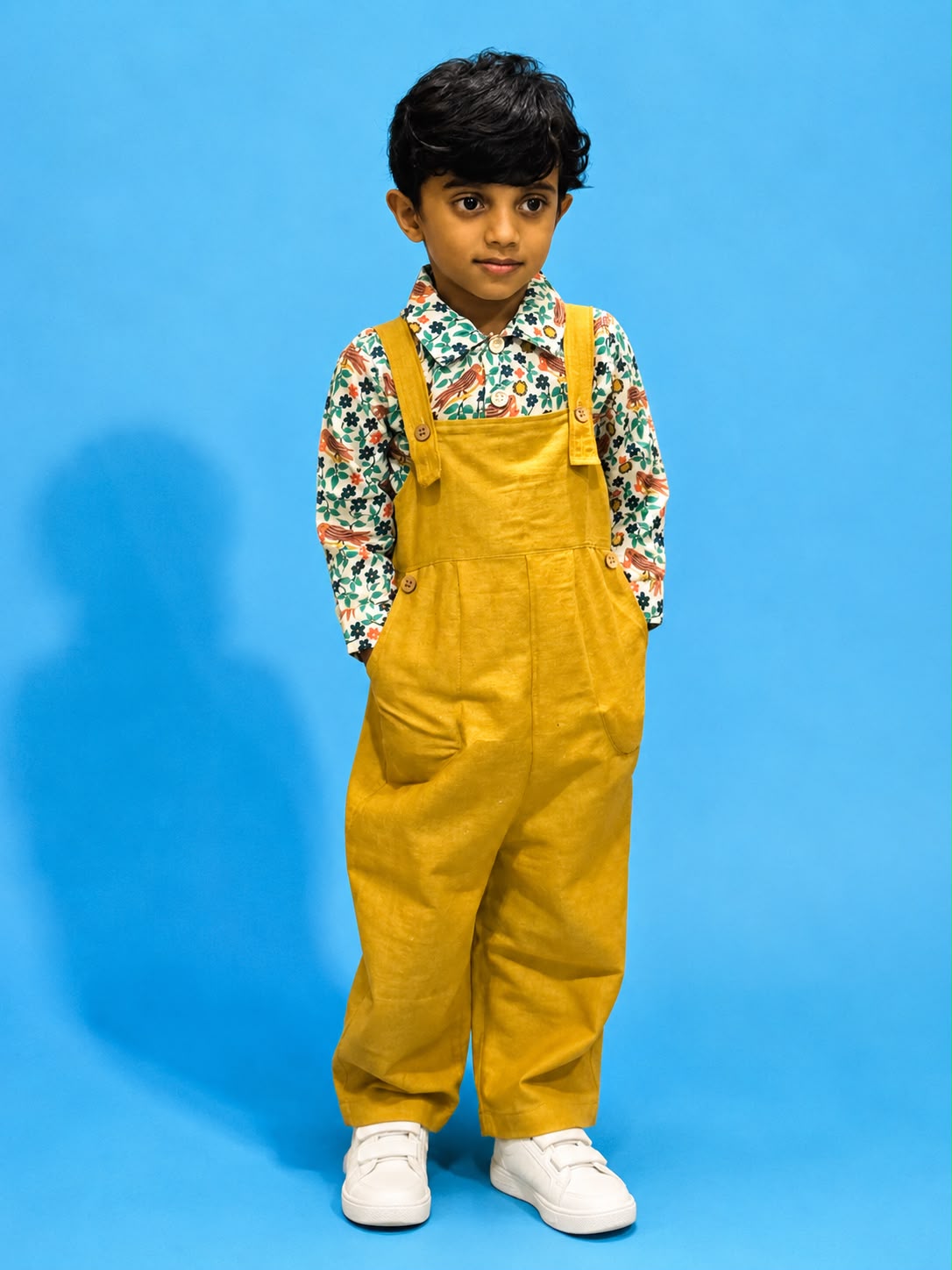 Boy standing in mustard khadi dungaree over floral print shirt and white sneakers.